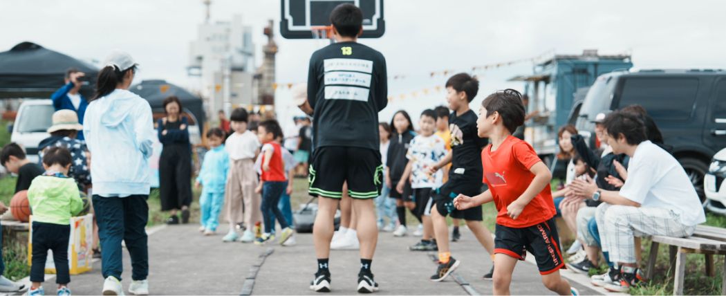 屋外のバスケットボールイベントで、大人や子供たちが楽しそうに見学している光景。