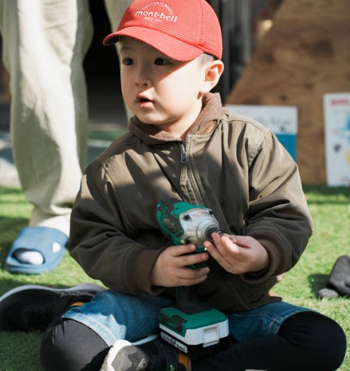 草の上に座り、赤いモンベル帽をかぶって電動ドライバーをいじる男の子。