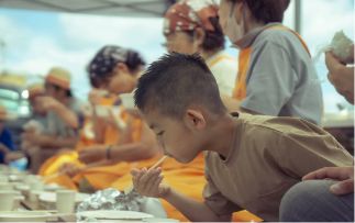 屋外で箸を使い食事をする少年と、並んで座る人々。