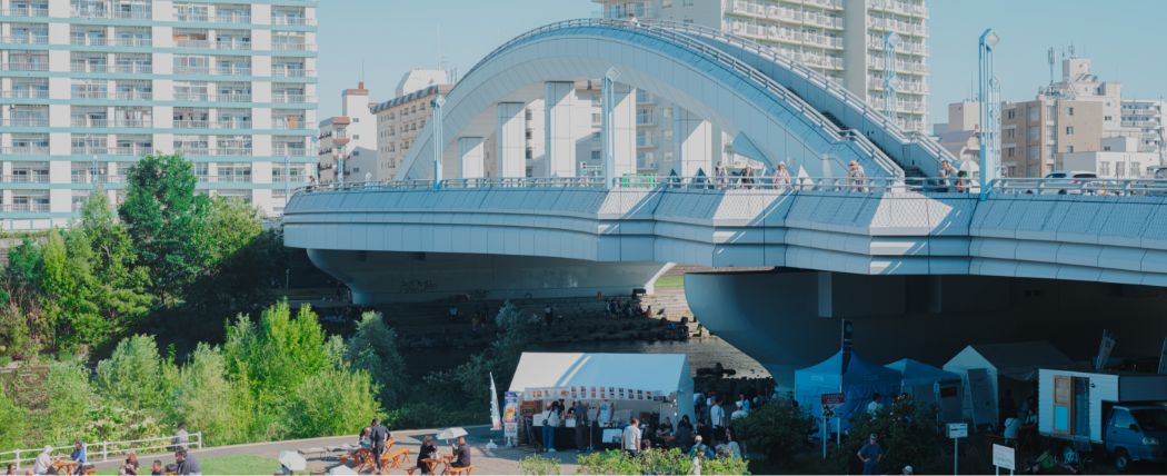 湾曲した橋の下で人々が賑わう川辺のマルシェ風景。