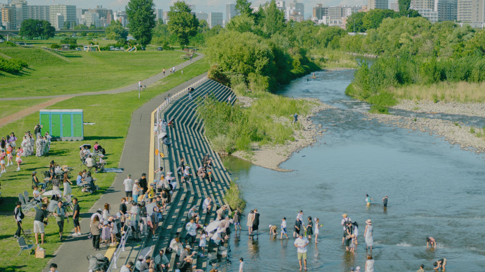 晴れた日に河川敷の公園でくつろぎ、川で水遊びをする人々。