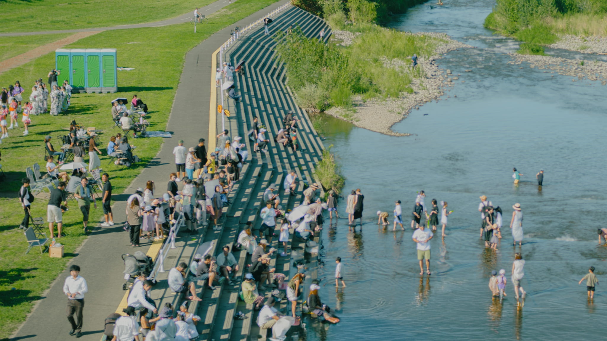 晴れた日に河川敷の公園でくつろぎ、川で水遊びをする人々。