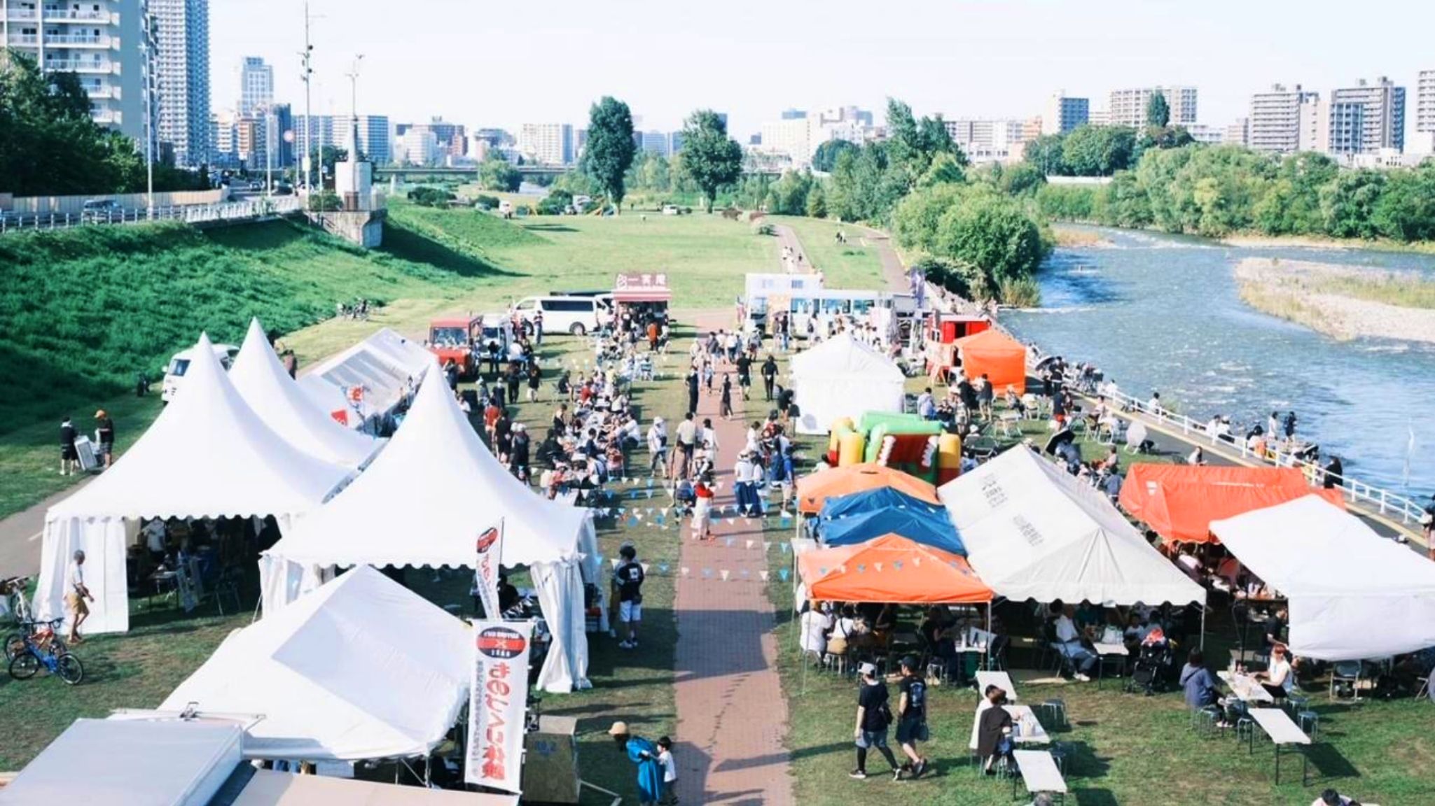都市の川沿い広場で、テントや屋台が立ち並び大勢の来場者で賑わうマルシェ。